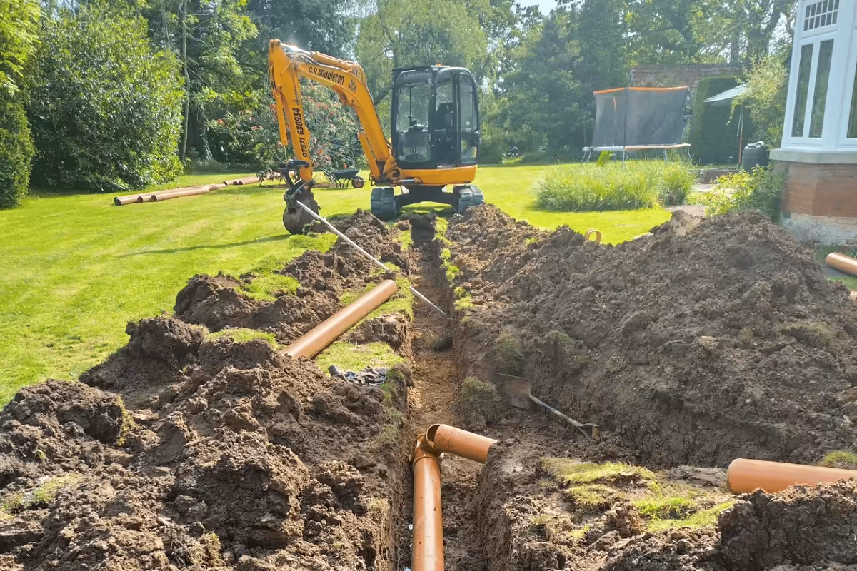 Excavator lowering drainage pipe into a trench across a lawn