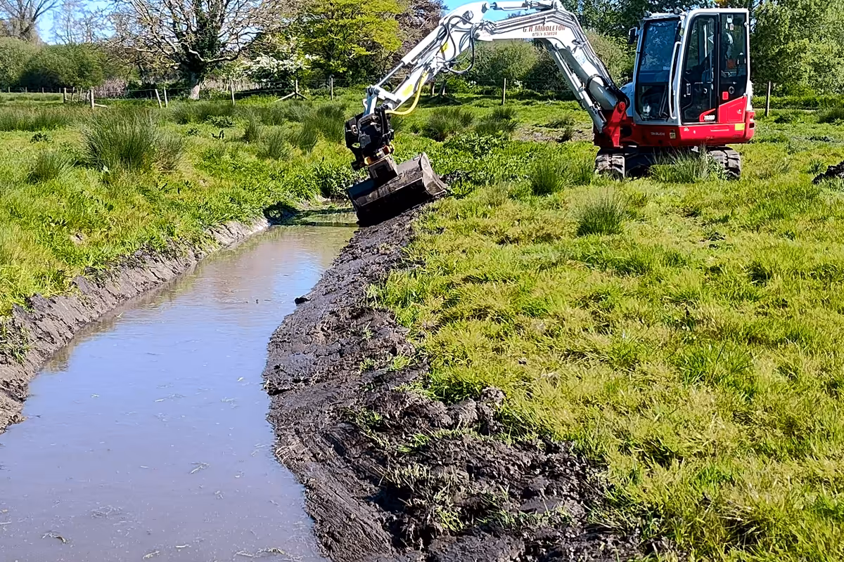 Excavator clearing a water-filled ditch through rough grassland