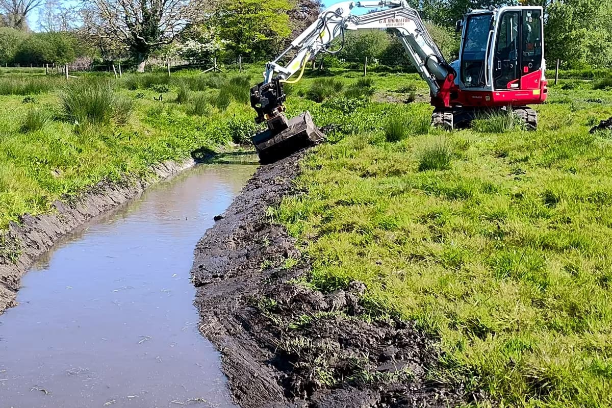 Excavator clearing a water-filled ditch through rough grassland