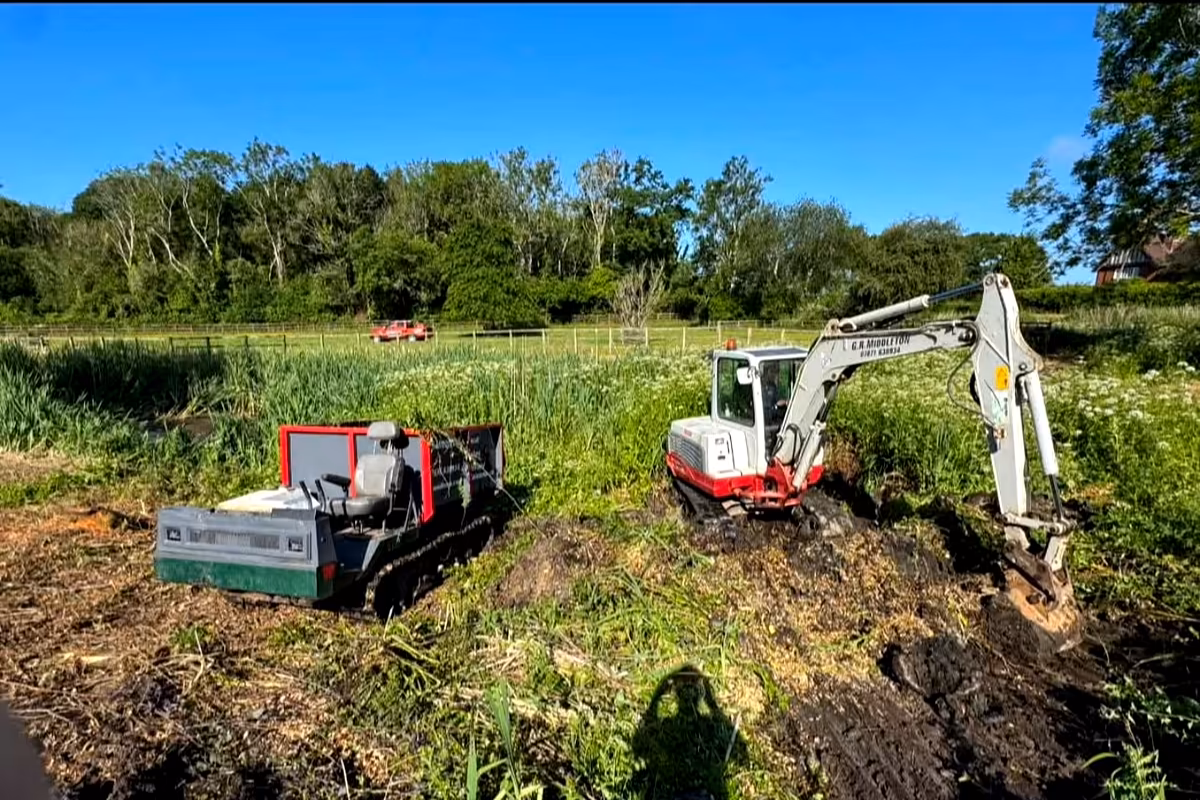 Excavator and tracked dumper at the overgrown edge of the pond