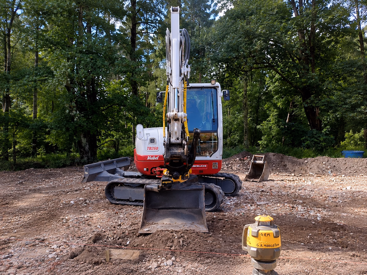 Gabriel's Takeuchi digger on a cleared plot, footings set out with string lines and a laser level in the foreground