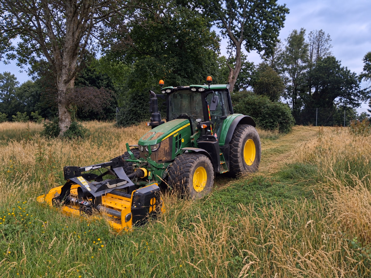 A tractor with a flail mower topping long grass under the trees, the cut ground behind it